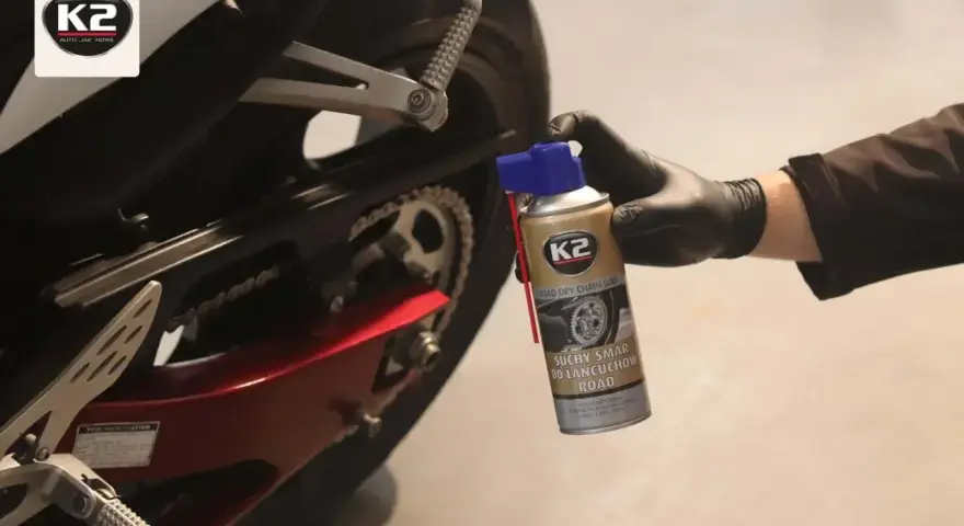 Ręka w czarnej rękawiczce aplikuje suchy smar do łańcucha motocyklowego K2. Idealny wybór, by zadbać o napęd.