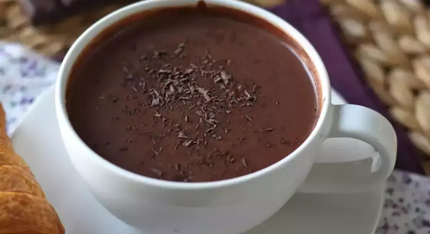 En vit mugg med varm choklad, toppad med chokladspån. En croissant ligger bredvid.