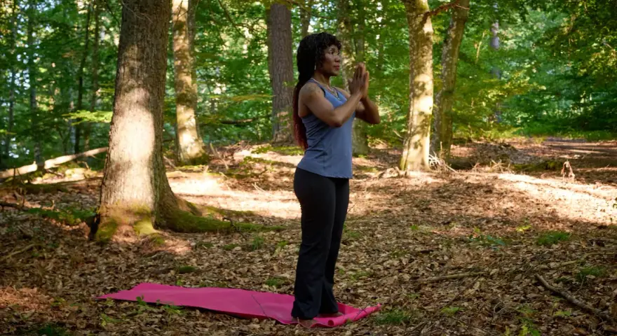 Kvinna i skogen på en yogamatta, med händerna i bön. En stund av avslappning och lugn för att minska stress.