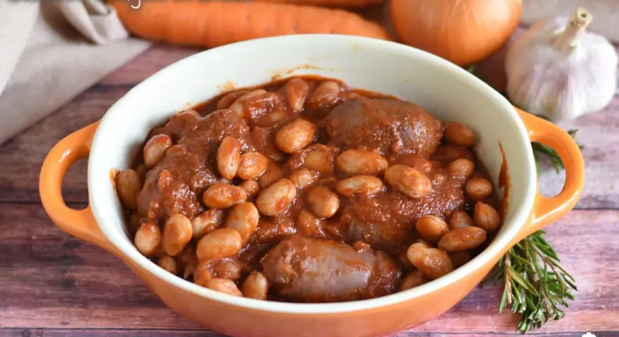 Piatto fumante di salsiccia e fagioli in umido, con carote, cipolla e aglio sullo sfondo.