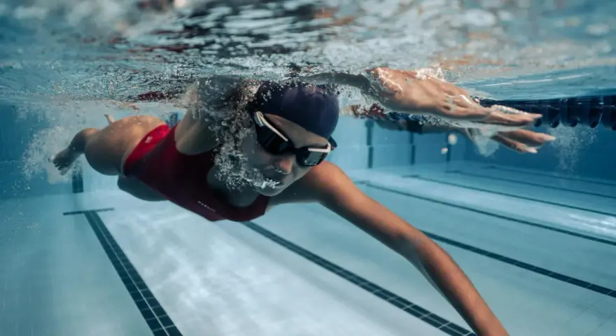 Une femme en maillot de bain rouge nage en crawl, lunettes de natation sur le nez. Sa **natation transformation physique femme** est visible sous l'eau.