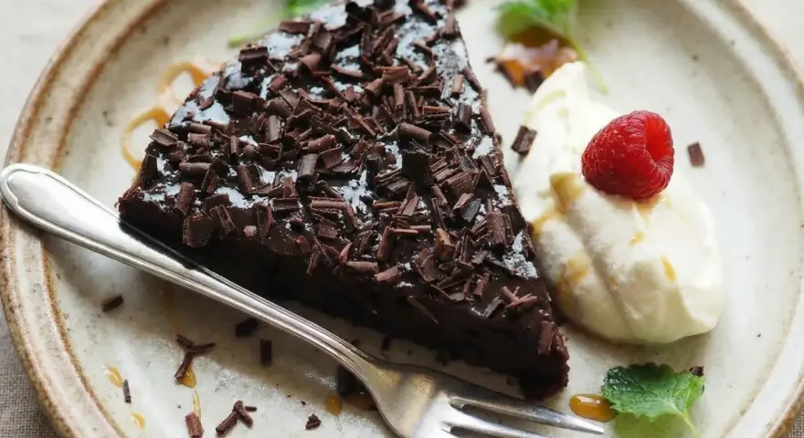 En bit glutenfri kladdkaka toppad med chokladspån, serverad med vispad grädde, hallon och mynta.