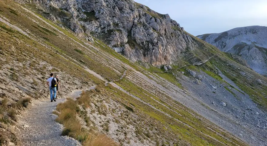 Due escursionisti percorrono un sentiero di trekking sul Gran Sasso, tra rocce e vegetazione alpina.