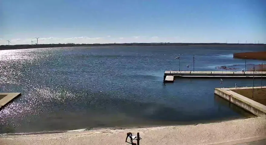 Widok z jeziorsko kamera na żywo. Na piaszczystej plaży dwie osoby, w tle spokojne jezioro, pomost i wiatraki.