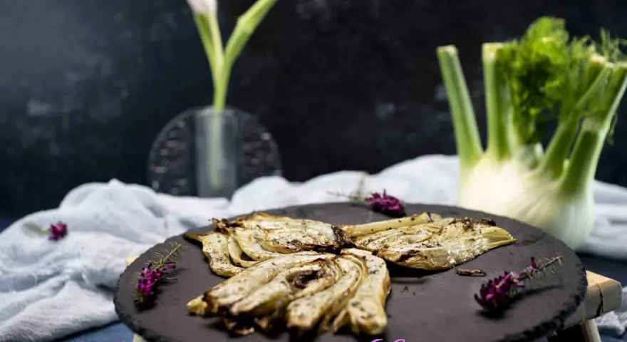 Finocchi croccanti e dorati, cotti nella friggitrice ad aria, serviti su una pietra scura con erbe aromatiche.