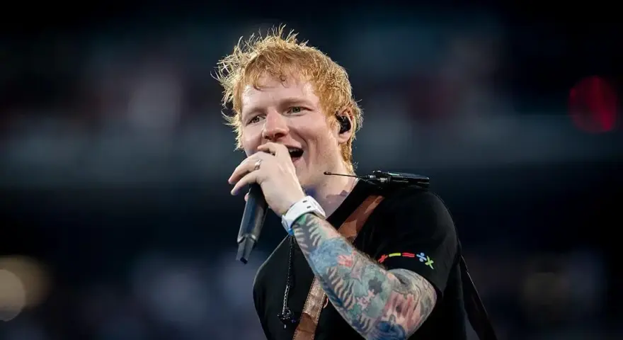 Ed Sheeran śpiewa z mikrofonem, jego ręka ozdobiona jest kolorowymi tatuażami.