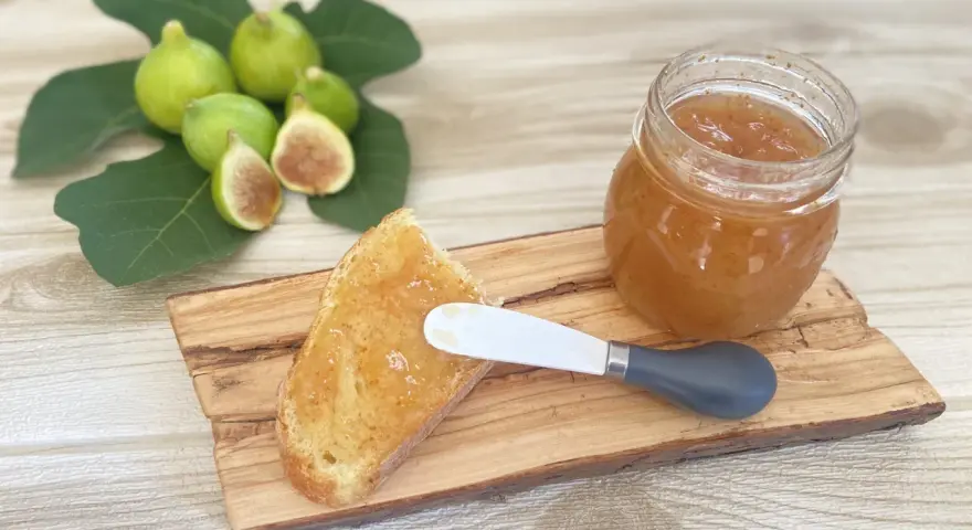 Fetta di pane con marmellata di fichi, accanto a un vasetto e a fichi freschi su un tagliere di legno.