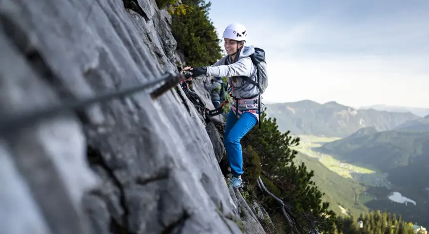 Kobieta w kasku i uprzęży pokonuje via ferrata, chwytając się stalowej liny. W tle rozciąga się malownicza górska dolina.