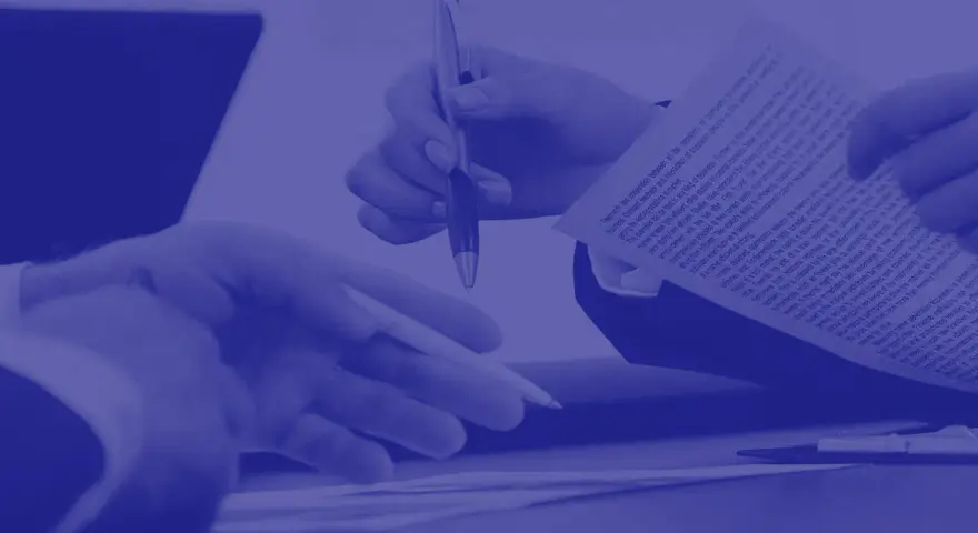 Dwie dłonie trzymają dokument, jedna z długopisem, druga wskazuje na tekst. To forma dokumentowa.