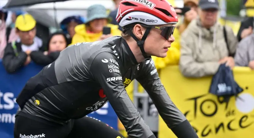 Trine Vingegaard, a cyclist in a grey and black racing suit, rides in the rain. Spectators watch from behind a yellow barrier with "Tour de France" branding.