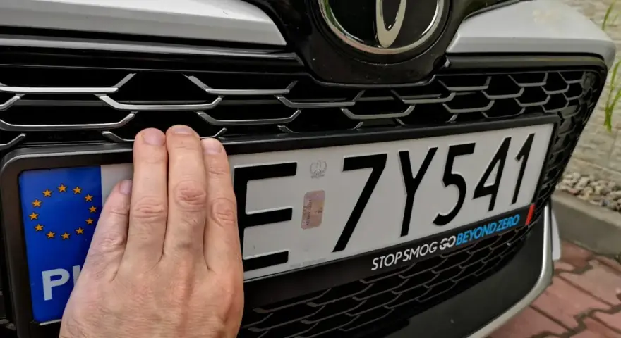 Ręka zasłania część tablicy rejestracyjnej. Upewnij się, że masz aktualne dokumenty, by uniknąć kary za nieprzerejestrowanie samochodu.