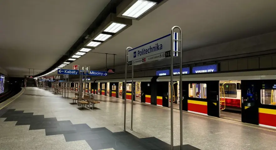 Pusta platforma stacji metra M1 Politechnika z pociągiem i tablicami kierunkowymi do Kabat i Młocin.
