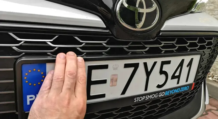 Dłoń zasłania część tablicy rejestracyjnej E 7Y541. Pod nią widoczny jest napis "STOP SMOG GO BEYOND ZERO".