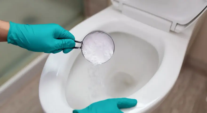 Hands in teal gloves pour baking soda into a toilet bowl, a method for how to unclog toilet without plunger.