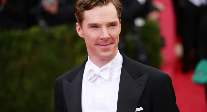Benedict Cumberbatch w czarnym smokingu i eleganckim białym krawacie, z uśmiechem pozuje na czerwonym dywanie.
