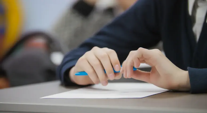 Uczeń pisze wypracowanie, przygotowując się do matury 2026. Na biurku biała kartka i niebieski długopis. Lektury obowiązkowe czekają.