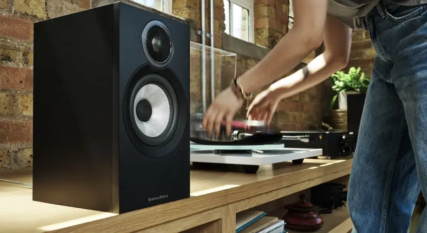 Czarny głośnik Bowers & Wilkins, jeden z najlepszych producentów głośników, obok gramofonu, na którym osoba kładzie płytę.