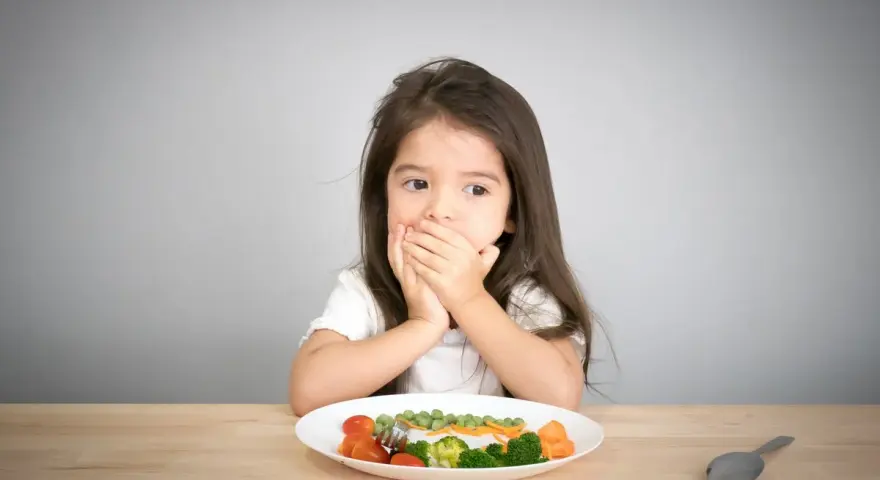Kleines Mädchen bedeckt Mund, schaut auf Teller mit Gemüse. Manche kinder essen schnell, aber dieses hier scheint zu überlegen.