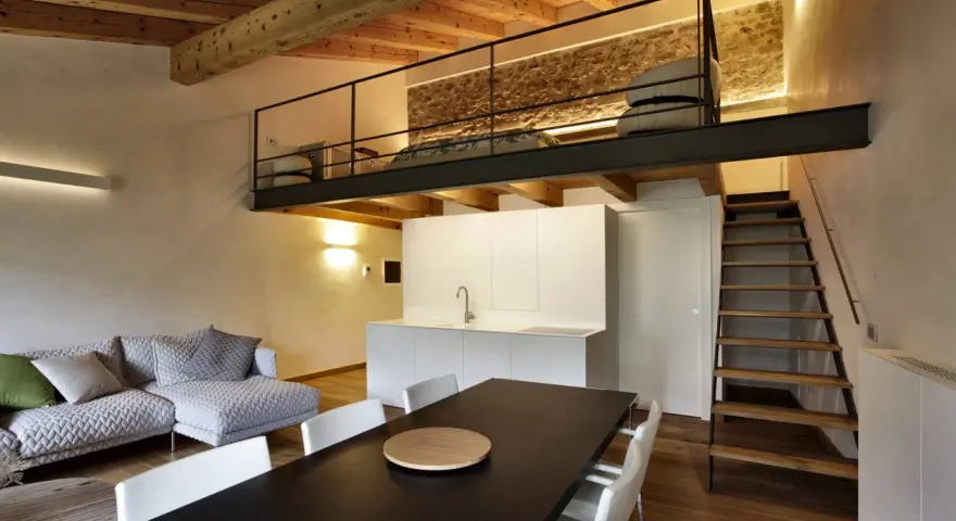 Modern living space with a loft bedroom accessible by stairs. The **soppalco soffitto basso** features a metal railing and a stone wall.