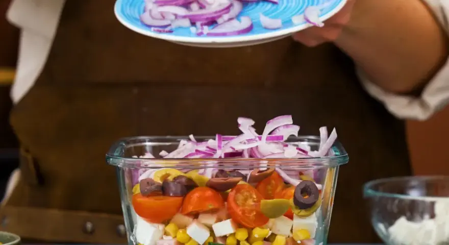 En person lägger rödlök på en pastasallad med kassler, majs, tomater, oliver och fetaost.