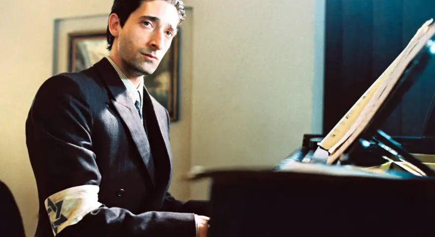 Adrien Brody jako Władysław Szpilman w filmie "Pianista" Romana Polańskiego, z opaską na ramieniu, siedzi przy fortepianie.