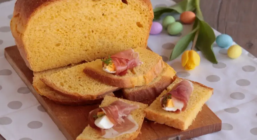 Fetta di torta al formaggio umbra ricetta originale, guarnita con uovo sodo e prosciutto crudo, su tagliere di legno, con uova colorate e tulipano.