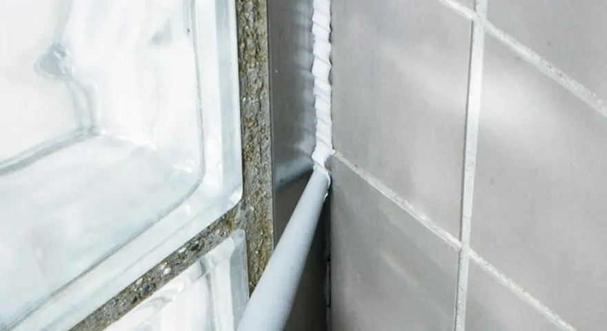 Applicator sprutar ny silikonfog längs en fog i badrummet, bredvid en duschvägg av glasblock.