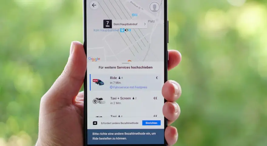 Hand hält Smartphone mit der besten Taxi App Deutschlands. Anzeige zeigt Fahrtoptionen nach Köln Hauptbahnhof.