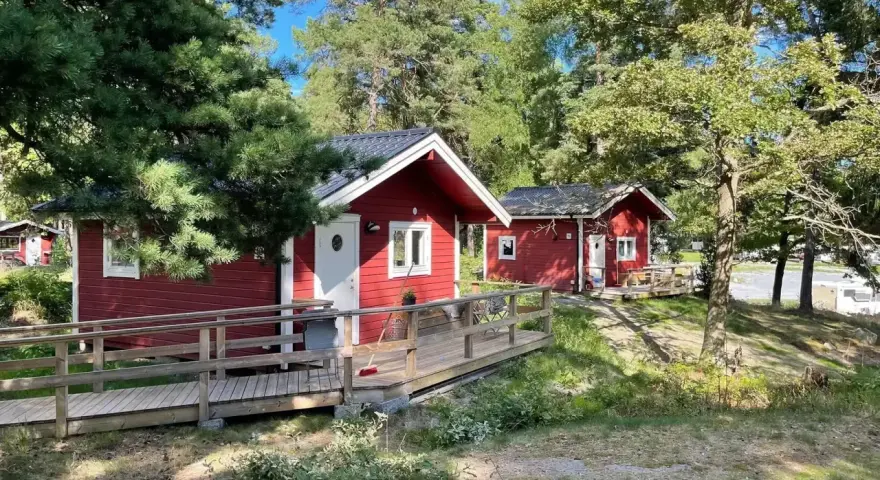 Röda stugor med träaltaner och ramper på Waxholms camping, omgivna av grönska och träd.