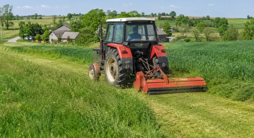 Czerwony traktor z mulczerem do c 330 kosi trawę na zielonym polu. W tle wiejskie domy i drzewa.