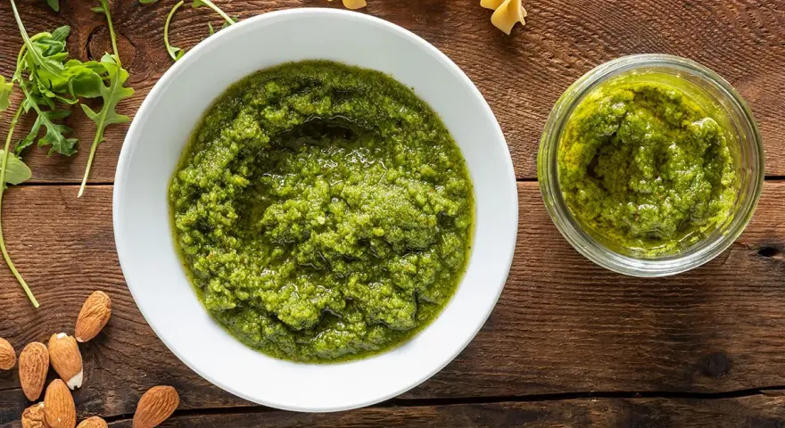 Pesto di rucola e mandorle in una ciotola bianca e un vasetto, con mandorle e rucola fresca intorno.