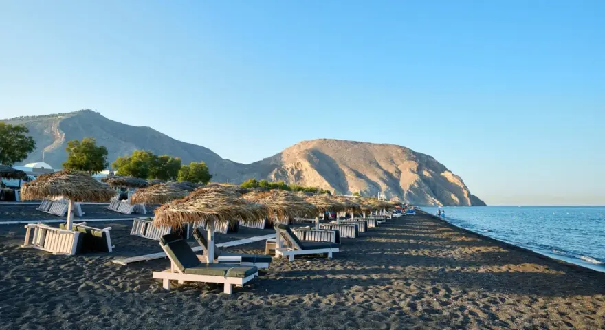 Czarne piaski i leżaki pod słomianymi parasolami na Santorini plażach. W tle góry i spokojne morze.