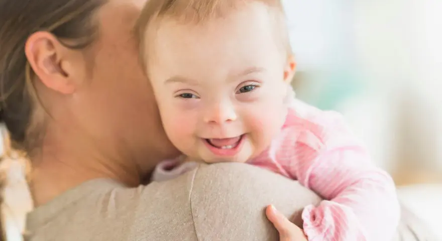 Ein lächelndes, leichtes Down-Syndrom Baby wird von seiner Mutter gehalten.