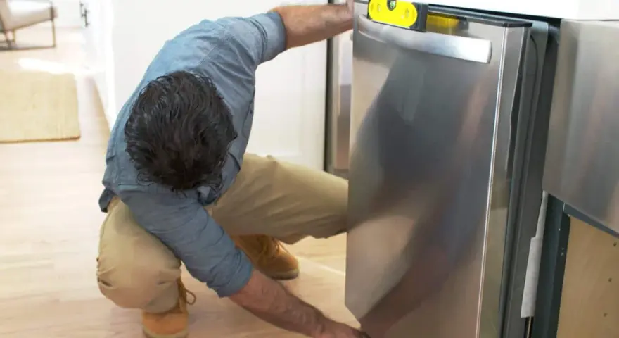 Man leveling a stainless steel dishwasher with a yellow bubble level, part of a guide on how to replace a dishwasher.