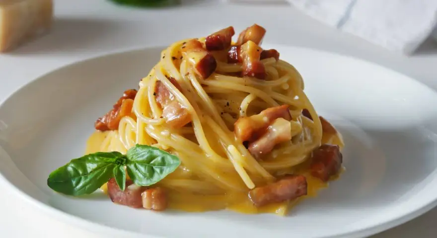 Pyszna carbonara z makaronem spaghetti, boczkiem i bazylią. W tle widać kawałek parmezanu, sugerując autentyczne carbonara skladniki.