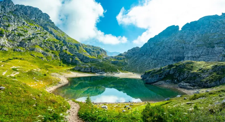 Sentiero panoramico tra le montagne, con un lago alpino dalle acque smeraldine che riflette il cielo azzurro e le nuvole bianche. Un perfetto percorso dolomiti.