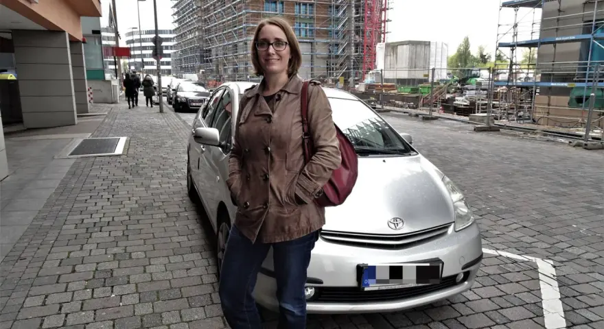 Frau mit brauner Lederjacke und roter Tasche steht neben silbernem Auto. Baustelle im Hintergrund. Sie ist free now fahrer.