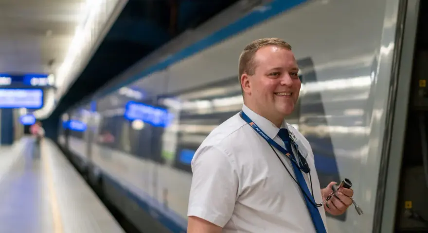 Uśmiechnięty konduktor PKP Intercity w białej koszuli i niebieskim krawacie stoi na peronie obok pociągu.
