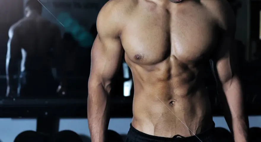 Muskulös man med synliga magmuskler i gym. En spegelbild av en annan person syns i bakgrunden. Frågan "hur deffar man" kan besvaras med denna fysik.