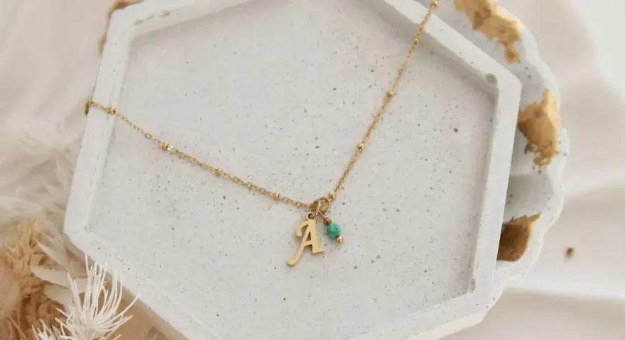 Złoty naszyjnik z literką "A" i turkusowym kamieniem, symbolizujący amulet na zajście w ciążę.