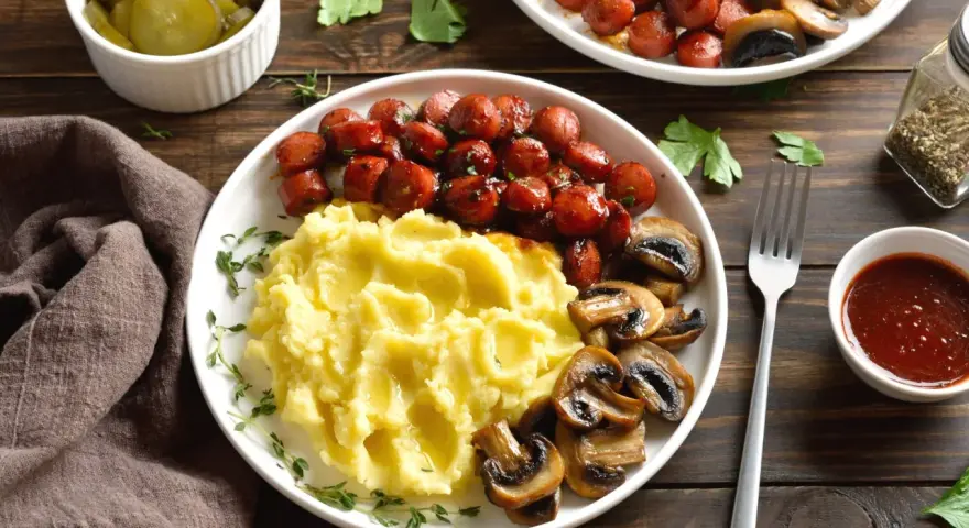 Talerz z purée ziemniaczanym, kiełbaskami i pieczarkami, obok miseczka z piklami i sosem.