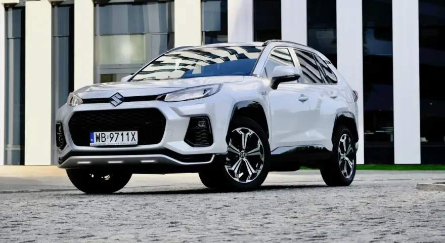 Biały Suzuki Across z czarnymi detalami stoi przed nowoczesnym budynkiem.