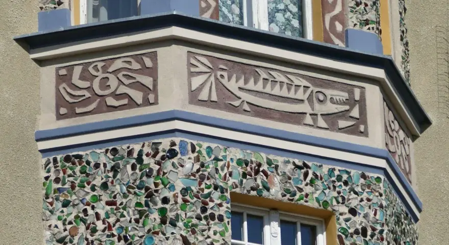 Sgraffito na fasadzie budynku: stylizowana ryba i abstrakcyjne wzory. Dolna część ozdobiona mozaiką z kolorowego szkła.