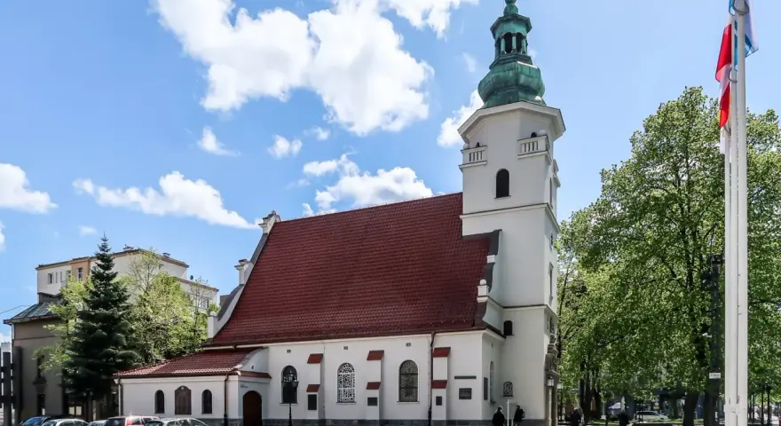 Biała bazylika pw. NMP Królowej Polski z czerwoną dachówką i zieloną wieżą, otoczona drzewami i budynkami.