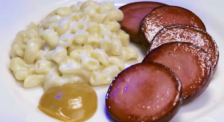 Stuvade makaroner snabbt vis med skivad falukorv och en klick senap på en vit tallrik.