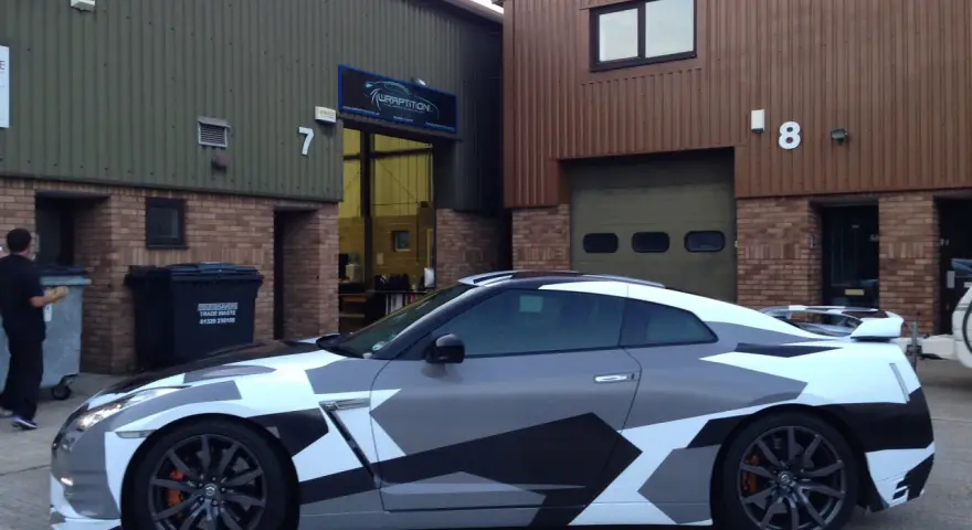 A Nissan GT-R with a striking geometric camouflage vinyl wrap for cars, parked outside a building with "WRAPTITION" signage.