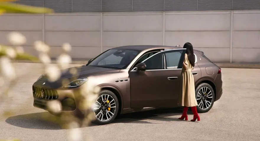 Elegancka kobieta w beżowej sukience i czerwonych kozakach zbliża się do brązowego Maserati SUV.