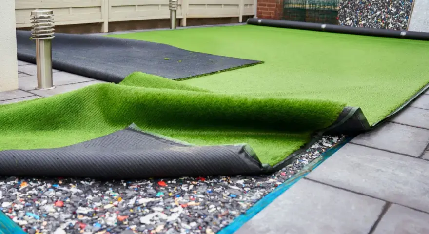 Instalación de césped artificial en terraza. Se ve el rollo de césped desplegado sobre una base de goma reciclada y baldosas.