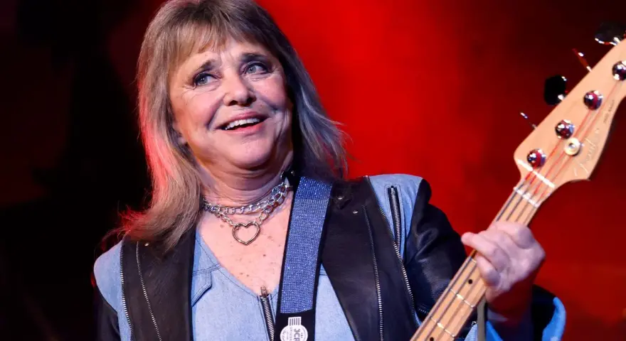Suzi Quatro, die Rock-Ikone, spielt Bassgitarre. Sie trägt eine Lederjacke und eine Kette mit Herzanhänger. Ihr Alter ist nicht ersichtlich, aber ihre Energie ist spürbar.