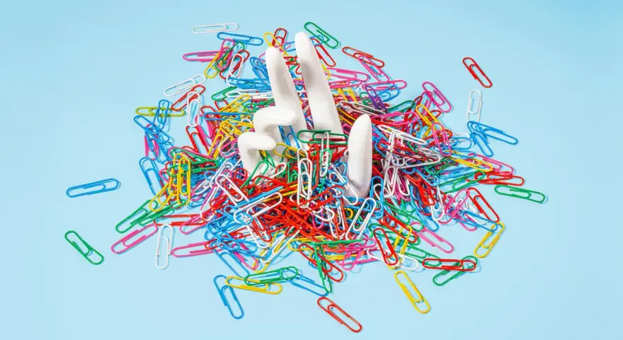 White hands emerge from a pile of colorful paperclips, symbolizing the struggle of how to stop being a workaholic and escape the daily grind.
